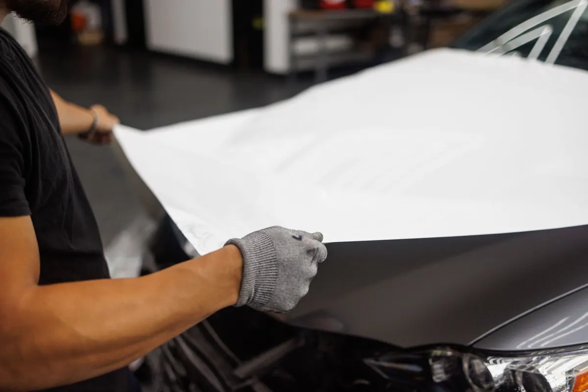 Hand mit Arbeitshandschuh legt eine weiße Folie glatt über eine Motorhaube