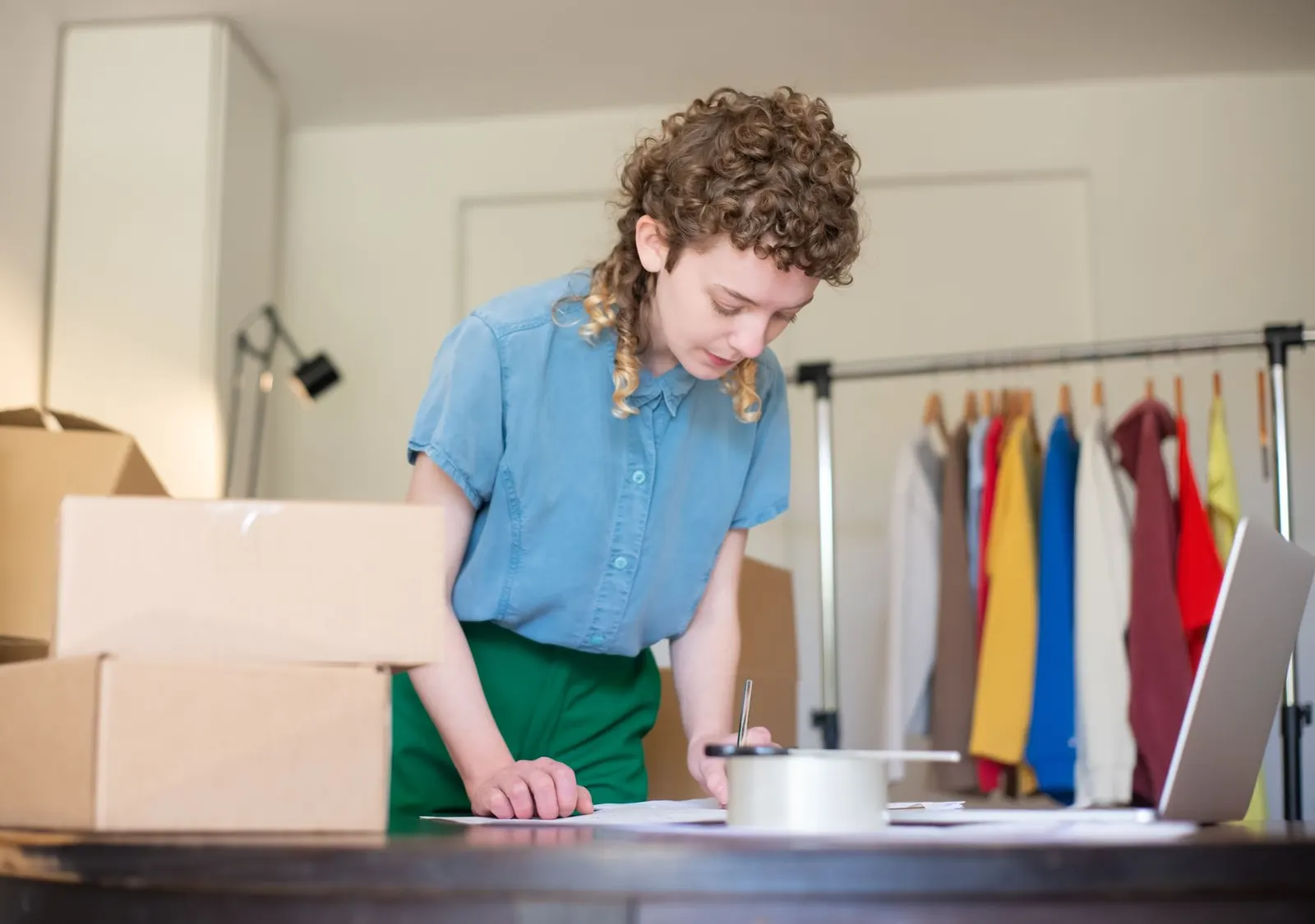 Inhaberin eines kleinen Betriebs arbeitet am Laptop zwischen Paketen und Kleiderständer